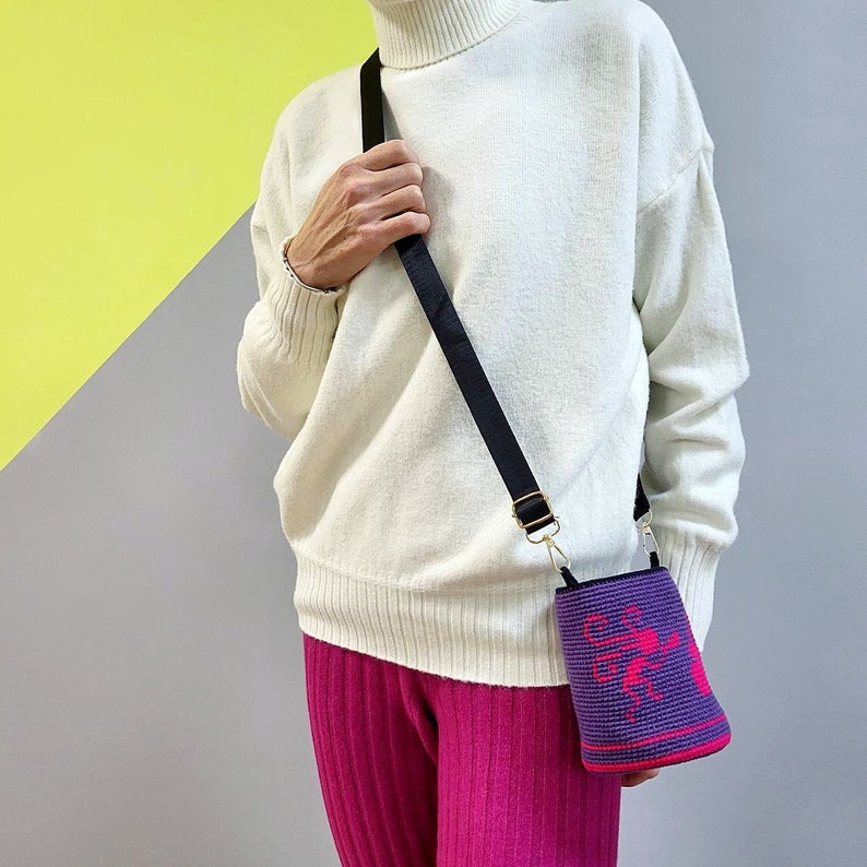 May include: A purple crocheted bag with a pink design hangs from a black strap on a person's shoulder. The bag is worn over a white sweater and pink pants.