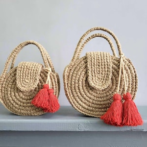 May include: Two round, tan-colored woven bags with top handles and a flap closure. Each bag features a textured, circular pattern and is decorated with bright red tassels attached to the handle. The bags are shown in two different sizes side by side.