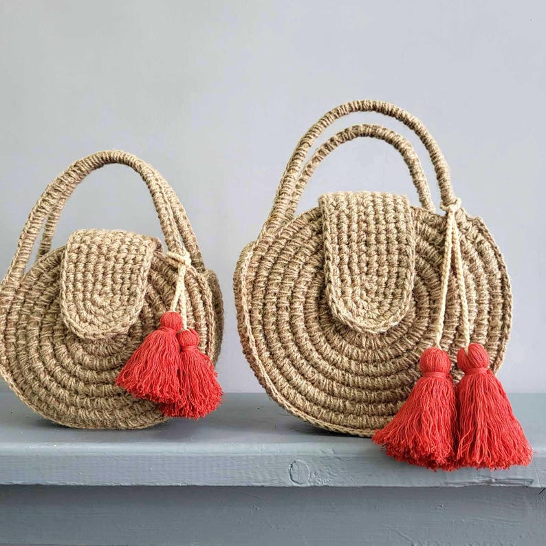 May include: Two round, tan-colored woven bags with top handles and a flap closure. Each bag features a textured, circular pattern and is decorated with bright red tassels attached to the handle. The bags are shown in two different sizes side by side.
