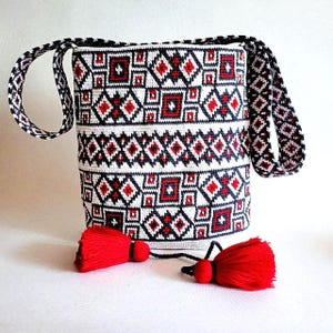 May include: White tote bag with a geometric pattern in red and navy blue. The bag has a matching strap and two red tassels hanging from the bottom. The design is a repeating pattern of diamonds and squares.