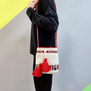 May include: White crocheted bucket bag with red and black embroidered floral details. The bag has a long strap and red tassels.