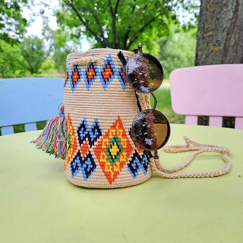 May include: A beige cylindrical woven bucket bag featuring a colorful geometric pattern in orange, blue, and yellow, with a multi-colored tassel on the side and a long braided strap, paired with round dark-tinted sunglasses resting on the bag.