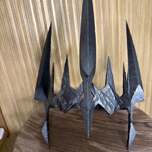 May include: A black metal crown with a pointed, jagged design. The crown has a sharp, angular shape and a textured surface.