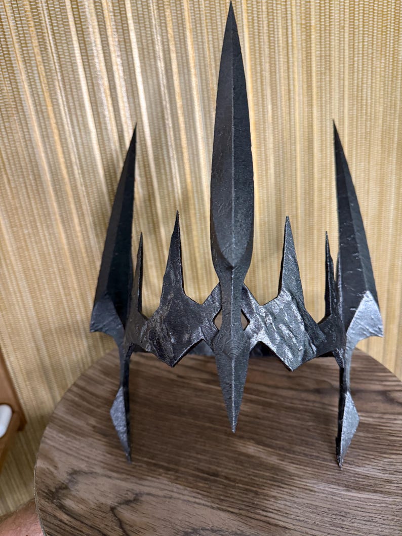 May include: A black metal crown with a pointed, jagged design. The crown has a sharp, angular shape and a textured surface.