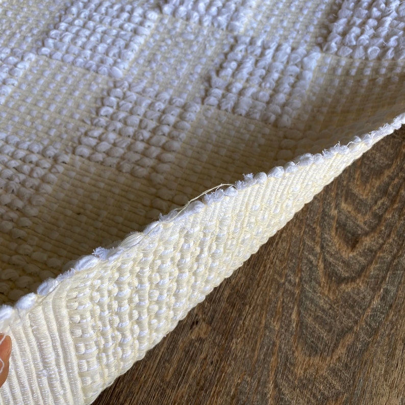 Pode incluir: Close-up de um tapete branco tecido com uma superf&iacute;cie texturizada. O tapete est&aacute; dobrado ao meio, revelando o lado de baixo da trama.