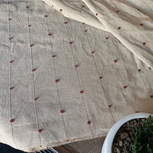 May include: Beige fabric with vertical lines of small, reddish-brown dots. The fabric has a textured appearance and is partially folded. The material appears to be natural, possibly linen or cotton, and is suitable for home decor or clothing.