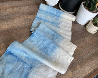 May include: A long, thin, blue and white linen table runner with a tie-dye pattern. The runner is folded in half and lying on a wooden surface.