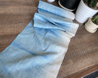 May include: A length of blue and white fabric, possibly linen or cotton, is rolled up at one end and unrolled at the other. The fabric has a watercolor-like effect, with darker blue hues blending into lighter shades.