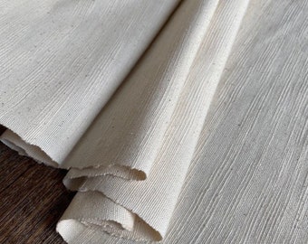Könnte beinhalten: Off-white Stoff mit einer strukturierten, gerippten Oberfläche. Der Stoff ist gefaltet, wodurch die rohe Kante sichtbar wird. Das Material scheint eine Naturfaser zu sein, möglicherweise Leinen oder Baumwolle, und eignet sich für Näh- oder Bastelprojekte.