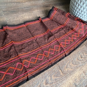 May include: A black fabric with a repeating geometric pattern in brown, orange, and pink. The fabric has a woven texture and is embroidered with a geometric design.