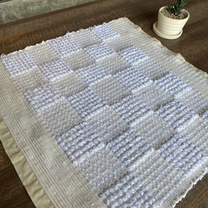 Pode incluir: Um tapete tecido branco e creme com um padr&atilde;o geom&eacute;trico de quadrados. O tapete tem uma superf&iacute;cie texturizada e uma borda de franjas.