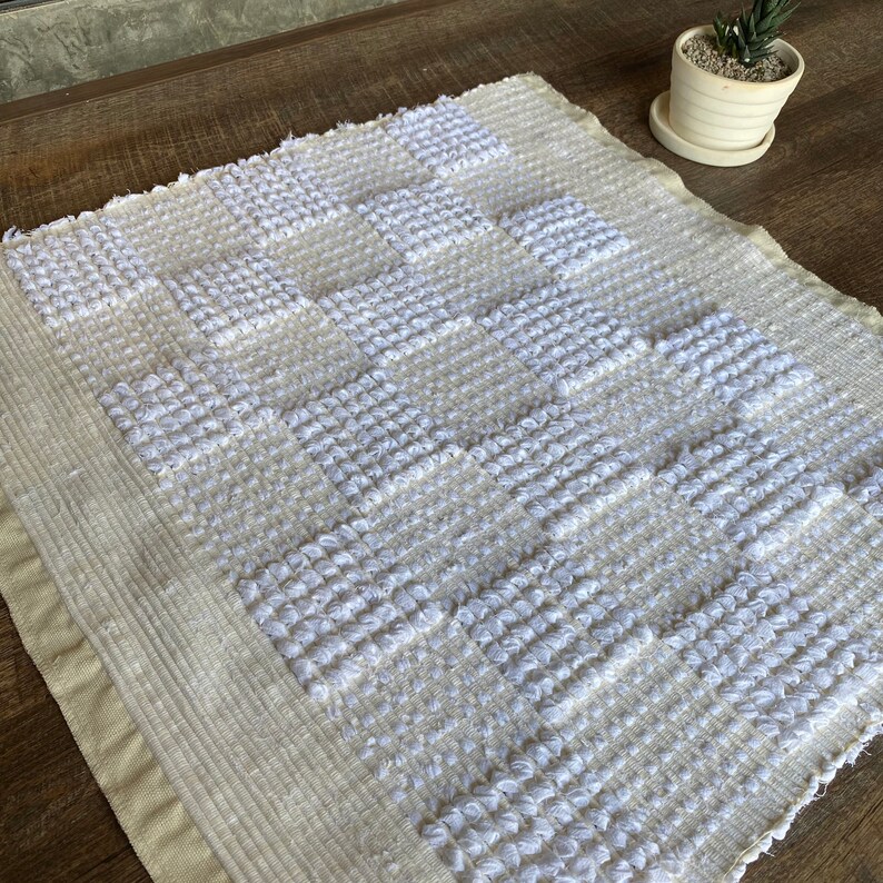 Pode incluir: Um tapete tecido branco e creme com um padr&atilde;o geom&eacute;trico de quadrados. O tapete tem uma superf&iacute;cie texturizada e uma borda de franjas.