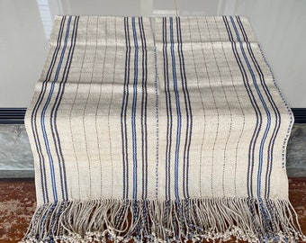 May include: A beige table runner with blue and brown stripes and fringe. The runner is made of a woven fabric and has a subtle texture.