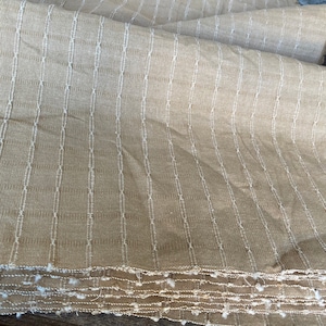 May include: A close-up of a beige fabric with white stripes. The fabric is folded and has a textured surface.