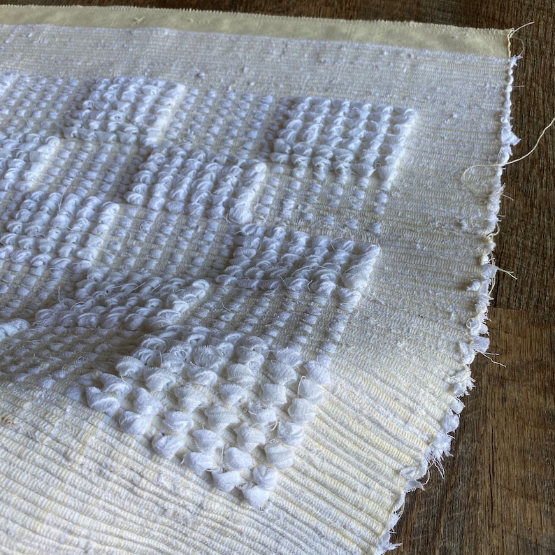 Pode incluir: Um tapete branco tecido com um padr&atilde;o texturizado em relevo. O tapete tem uma borda de franja e &eacute; feito de fibras naturais.