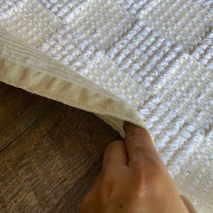 Pode incluir: Close-up de um tapete de banho texturizado branco e creme com uma borda bege. O tapete est&aacute; dobrado e uma m&atilde;o est&aacute; segurando a borda do tapete.