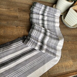 May include: A black and white striped fabric runner with a fringed edge. The runner is folded in half and lying on a wooden surface.
