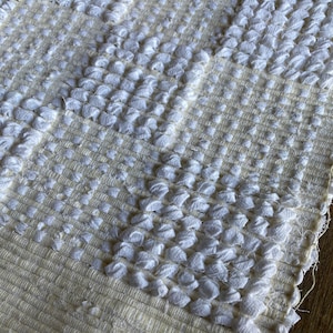 Pode incluir: Close-up de um tapete tecido texturizado branco e creme com um padr&atilde;o em relevo. O tapete tem uma textura macia e fofa.