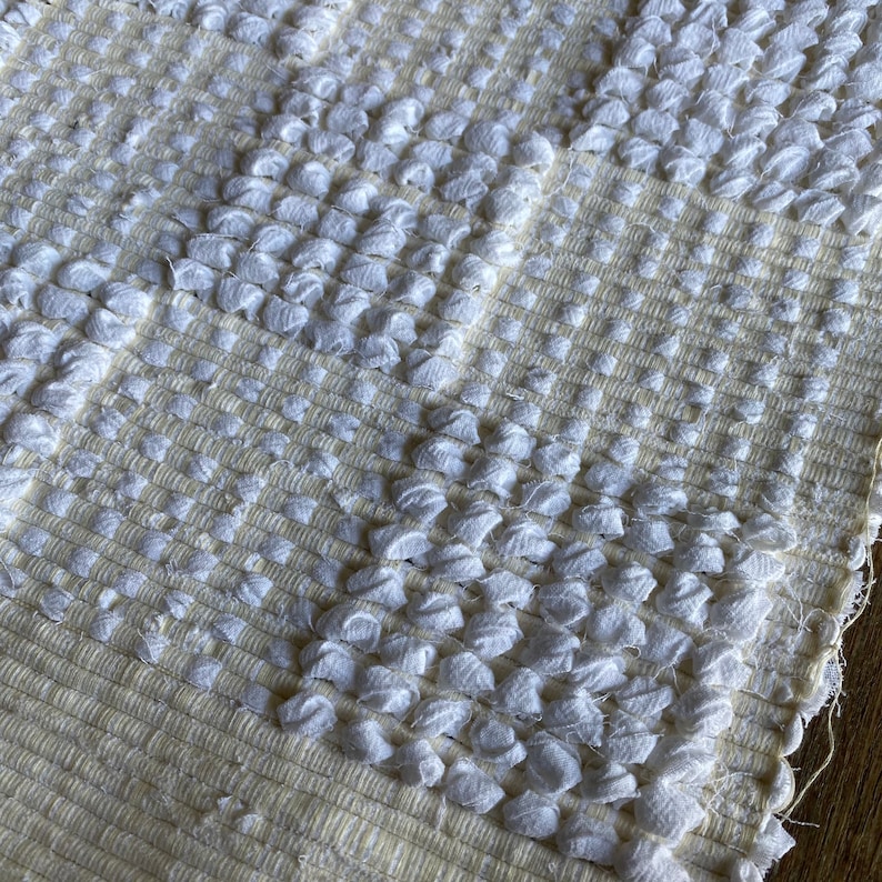 Pode incluir: Close-up de um tapete tecido texturizado branco e creme com um padr&atilde;o em relevo. O tapete tem uma textura macia e fofa.