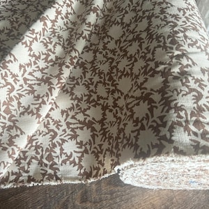 May include: Cream-colored fabric with a repeating pattern of brown floral shapes. The fabric is folded and rolled, revealing the raw edge of the material. The design is reminiscent of a botanical print, suitable for home decor or apparel.
