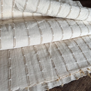 Puede incluir: Primer plano de una tela blanca rota doblada con filas verticales de costuras beige. La tela tiene un aspecto texturizado y un borde sin rematar. Las costuras crean un patrón lineal sutil. La tela parece ser una fibra natural.