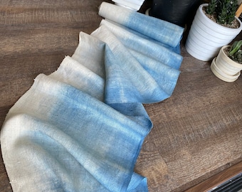 May include: A long, thin piece of fabric with a gradient of blue dye, fading to white at one end. The fabric is folded and draped on a wooden surface.