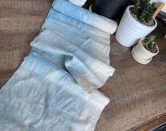 May include: A light blue and white ombre fabric runner with a textured surface. The runner is folded and draped on a wooden surface.