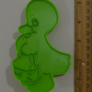 Puede incluir: Cortador de galletas de plástico verde en forma de pato de dibujos animados con una cabeza grande y un cuerpo pequeño. El pato tiene un pico grande, ojos pequeños y una cola pequeña. El cortador de galletas mide aproximadamente 7,5 cm de alto.