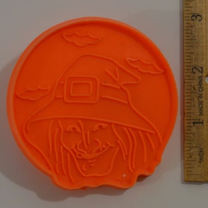 May include: Orange plastic cookie cutter shaped like a witch with a pointed hat and a wide grin. The cutter is approximately 5 cm in diameter.