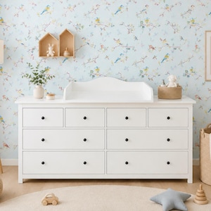 Op de afbeelding: Een witte houten ladekast met acht lades voorzien van kleine zwarte ronde knoppen en een bijpassend wit aankleedblad. De kast heeft een rechthoekig frame met een platte basis en een gebogen achterrand op het bovenoppervlak.