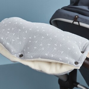 Könnte beinhalten: Graue Kinderwagen-Lenkerbezüge mit einem weißen Löwenzahnmuster und einem weißen Fleece-Futter. Die Bezüge haben einen Druckknopfverschluss und ein braunes Leder-Etikett mit dem aufgedruckten Wort "Kooduu".
