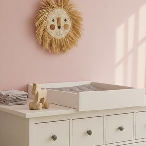 Peut inclure: Une scène de chambre d'enfant avec une table à langer blanche, un insert rembourré gris, un cheval à bascule en bois et des vêtements gris pliés. Une décoration murale en tête de lion tissé est accrochée au mur rose.