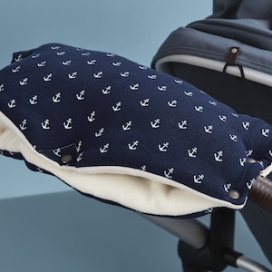 Könnte beinhalten: Marineblauer Kinderwagen-Handwärmer mit weißem Anker-Muster. Die Innenseite ist aus weichem, cremefarbenem Material. Der Handwärmer wird mit Druckknöpfen befestigt und soll die Hände warm halten. Der Kinderwagen-Griff ist braun.