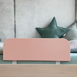 Può includere: Testiera rosa pallido fissata a un telaio letto in legno chiaro. Il letto è fatto con un lenzuolo verde chiaro e una coperta fantasia. Cuscini a forma di stella verdi e grigi sono appoggiati a un muro di cemento.