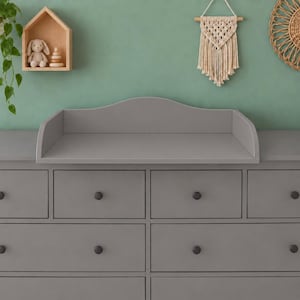 Op de afbeelding: Een grijze houten ladekast met zes lades voorzien van eenvoudige ronde knoppen en een bijpassend aankleedblad met een gebogen achterpaneel. De ladekast biedt een vlak oppervlak voor babyverzorging en is afgewerkt in een matte grijze lak.