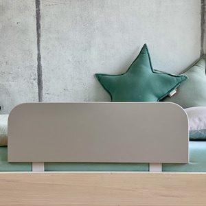 Könnte beinhalten: Ein hellgraues Bettgitter mit abgerundeten Kanten, befestigt an einem Holzbettgestell. Das Bett hat ein hellgrünes Laken und ein weißes Kissen mit geometrischem Muster. Zwei grüne sternförmige Kissen liegen auf dem Bett hinter dem Gitter.