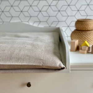 Op de afbeelding: Een witte commode met een beige corduroy verschoonmat. De commode heeft een lade met een geborsteld nikkel knopje. Er liggen houten blokken en een geweven mand op de commode.