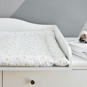 Puede incluir: Mesa cambiador blanca con una colchoneta cambiador blanca con un patrón amarillo de pequeñas zanahorias. Una manta blanca con un patrón amarillo de zanahorias está doblada sobre la mesa con un pequeño cordero de peluche blanco encima.