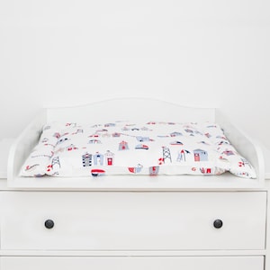 Puede incluir: Mesa cambiador blanca con una colchoneta cambiador blanca con un estampado náutico de casas y barcos rojos, azules y blancos. La mesa cambiador tiene tres cajones.