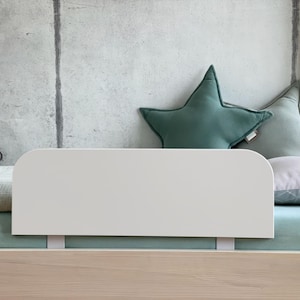 Può includere: Sponda letto bianca con bordi arrotondati, fissata a una struttura letto in legno. Il letto ha un lenzuolo verde chiaro e un cuscino bianco con un cuscino a forma di stella verde sopra.