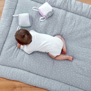 Könnte beinhalten: Ein Baby in einem weißen Strampler krabbelt auf einer grau-weißen Spielmatte. Auf der Matte liegen drei bunte Stoffbausteine.
