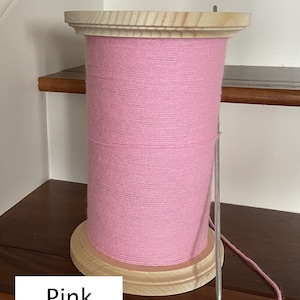 Puede incluir: Una gran bobina de hilo rosa con base y tapa de madera. Una aguja de metal está insertada en el hilo, con un hilo de hilo rosa que se extiende desde él. La palabra "Pink" se muestra en un rectángulo blanco en la parte inferior.