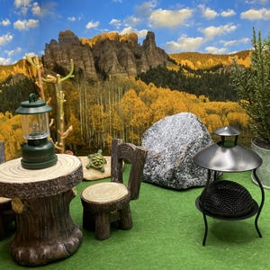 May include: Miniature outdoor scene with a rustic wooden table and chairs, a green lantern, and a small metal fire pit. A large rock and a potted plant are also present, set against a backdrop of a mountain landscape.