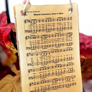 May include: A rectangular, vintage-style ornament featuring sheet music with the title "Blessed Assurance: Jesus Is Mine." The aged paper has visible musical notes and lyrics. A natural twine string is attached for hanging. The background includes fall foliage.