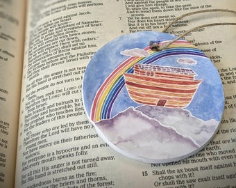 May include: Round, watercolor-style ornament featuring Noah's Ark, a rainbow, and mountains against a blue sky. The ornament has a twine loop for hanging. The theme is religious.