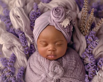 May include: A newborn baby wearing a purple turban with a flower on top, sleeping peacefully surrounded by purple flowers.