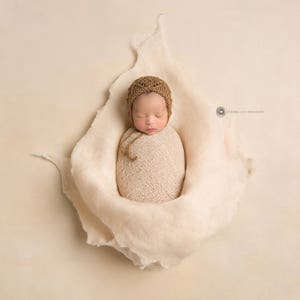May include: A newborn baby is swaddled in a cream-colored blanket and wearing a brown knitted hat. The baby is sleeping peacefully on a cream-colored background.