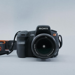 May include: Black Sony α200 DSLR camera with a lens and attached strap. The lens has a 0.38m/1.3ft macro setting. The strap is black with orange accents. The camera is on a light gray background.