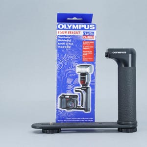May include: An Olympus Flash Bracket with a black handle and base, and a blue box with the product name and a diagram of the bracket with a camera and flash attached. The bracket is designed to hold a camera and flash.