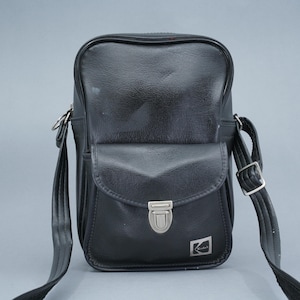 Può includere: Borsa per fotocamera in pelle nera con patta frontale e chiusura argentata. La borsa ha una tracolla e un piccolo logo Kodak sul davanti. Progettata per trasportare una fotocamera e accessori.